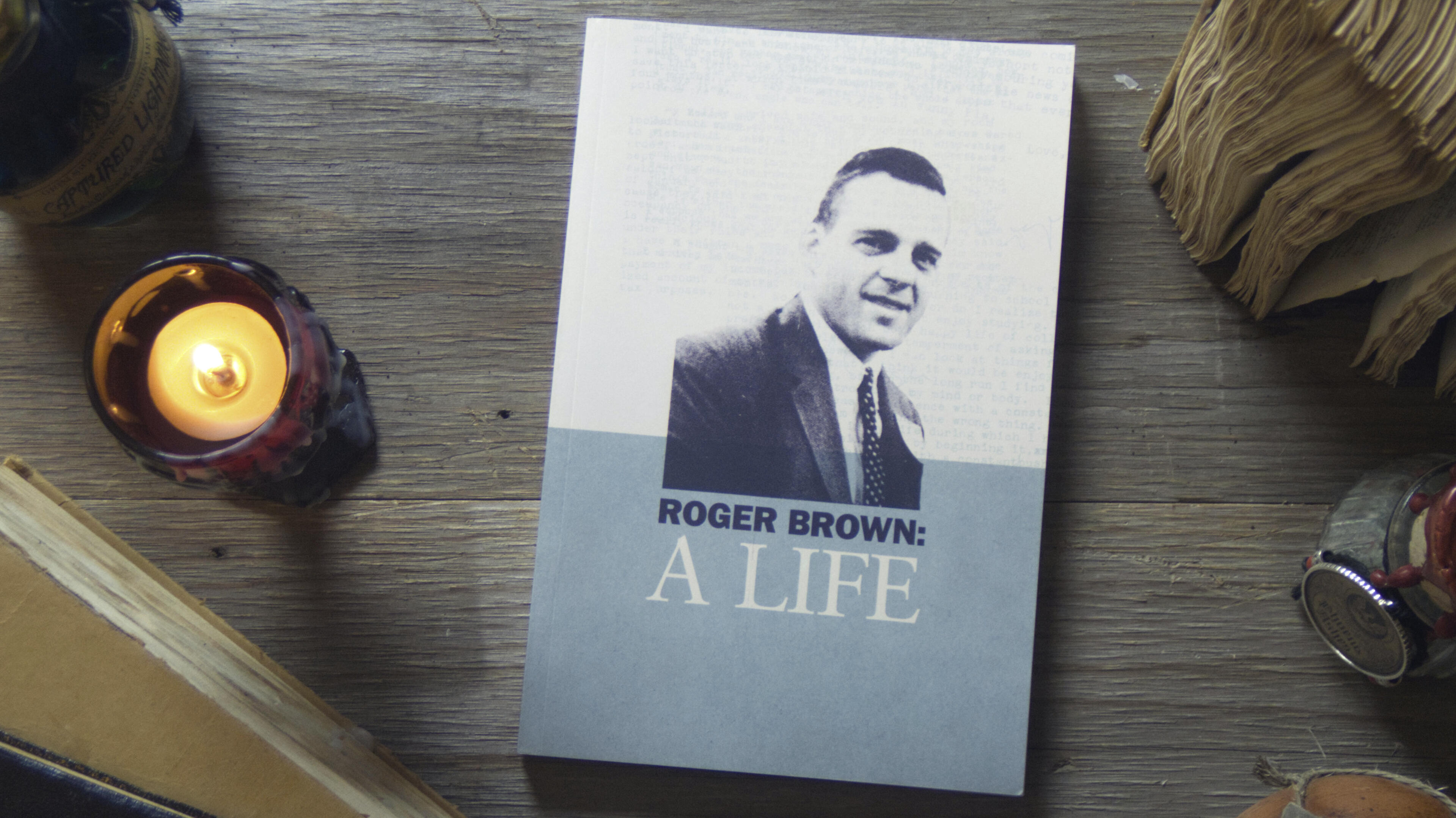 Roger Brown (Front) On top of a wooden table, a scattering of old books and gothic candles set the scene. In the center is a fresh paperback copy of Roger Brown: A Life. It is a dignified design with gentle colors suggesting the rich Chicago history in its pages.